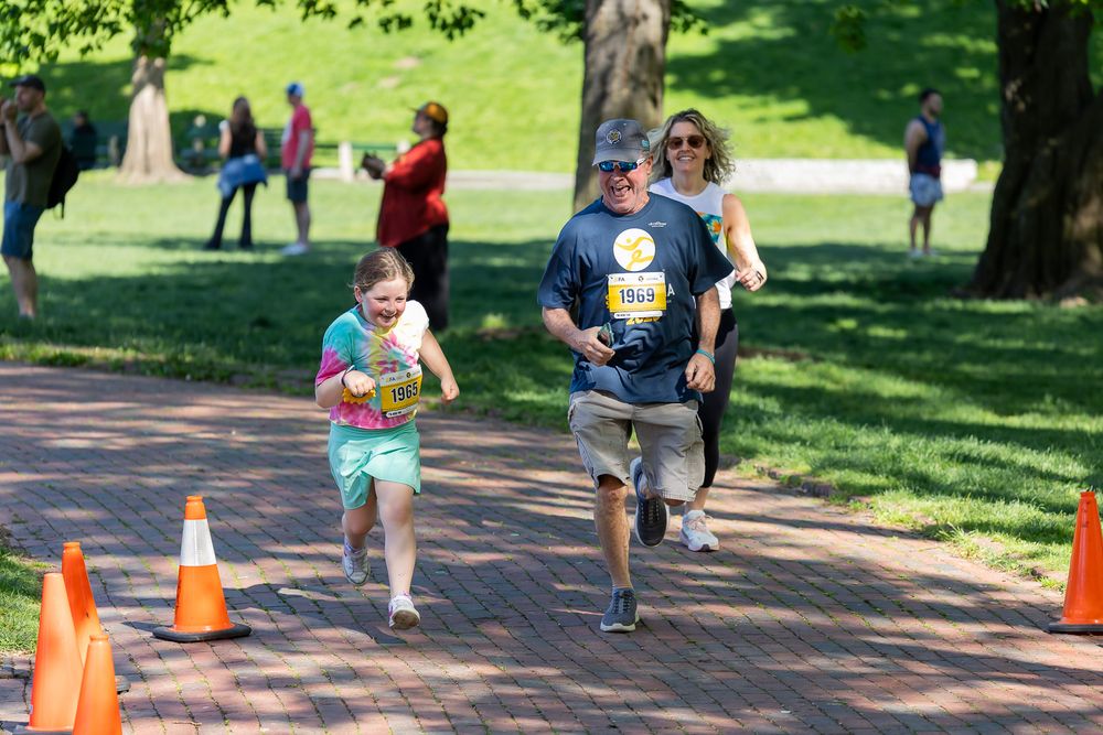 Children and adults running on a brick path in a park during an outdoor charity fun run event on a sunny day.