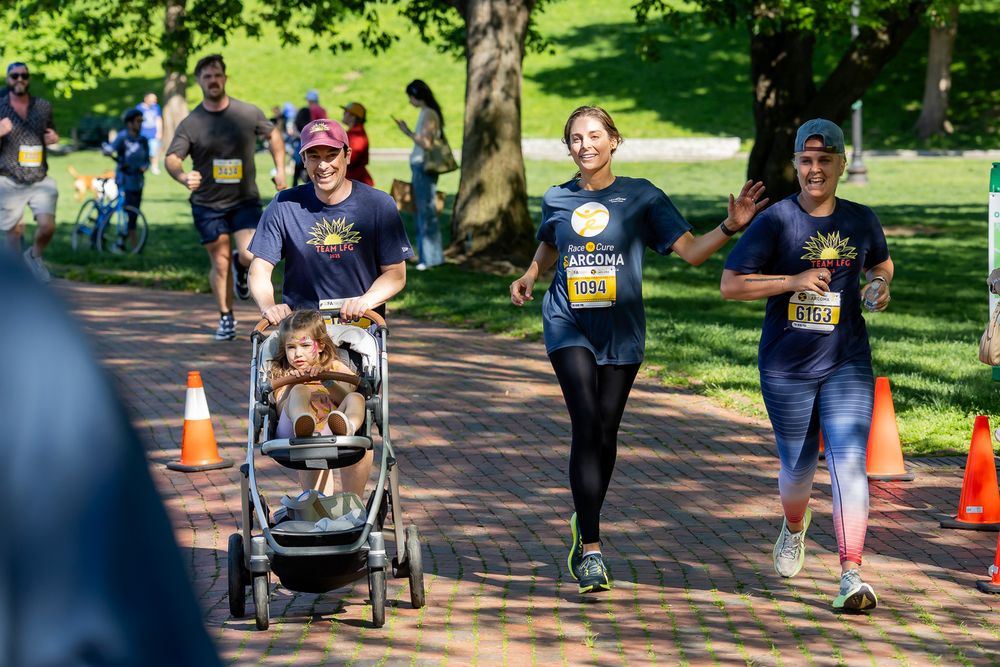 A group of runners in matching shirts jog through a park, one pushing a stroller with a child during a race.
