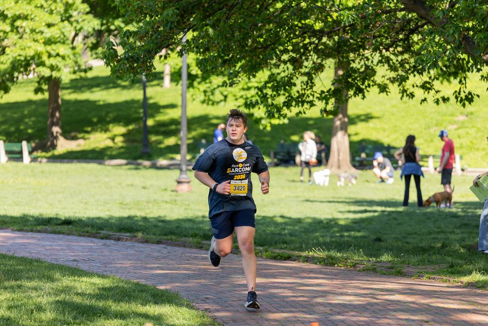 A man in a black shirt runs along a brick path through a sunny green park during a race event.