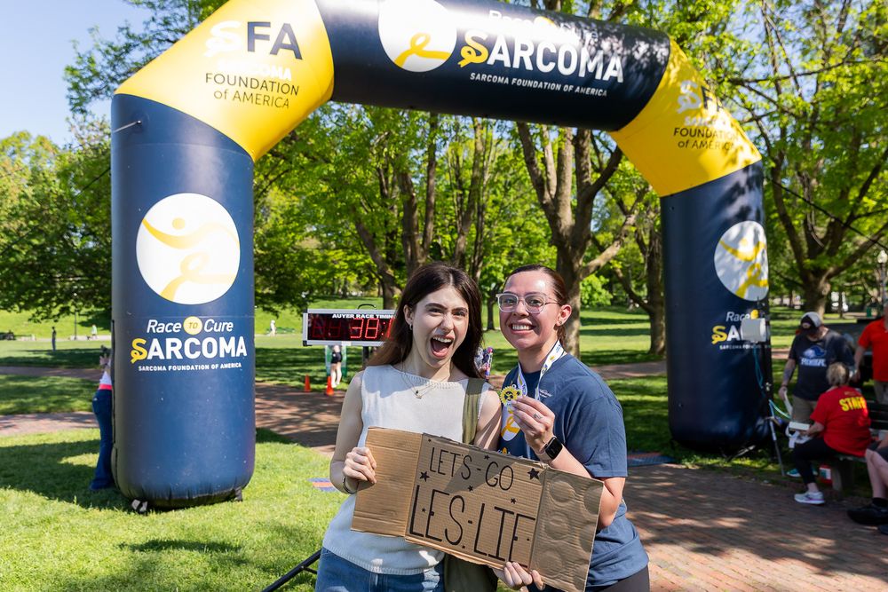 Two smiling participants pose under the Sarcoma Foundation of America Race to Cure arch holding a sign.