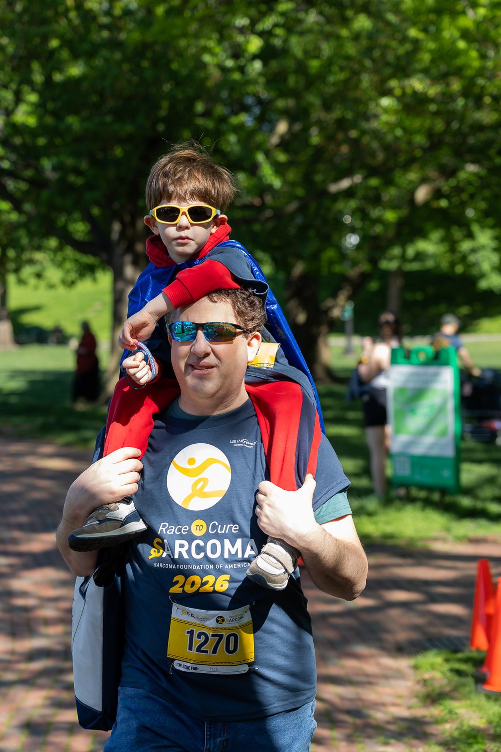Man carrying child on shoulders at Race to Cure Sarcoma event, child wearing superhero cape and yellow sunglasses.