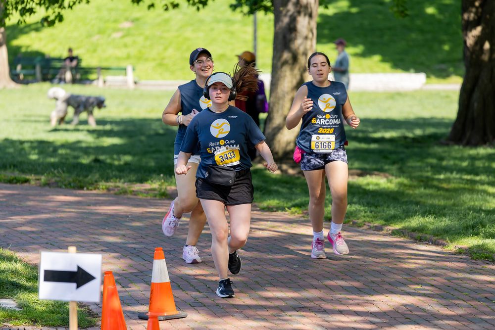 Three runners compete in an outdoor road race through a sunny park, wearing race bibs and athletic gear.