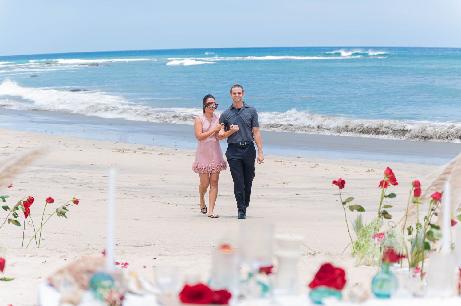 Couple walking hand-in-hand on a beach with red roses and candles in the foreground during a romantic proposal setup.