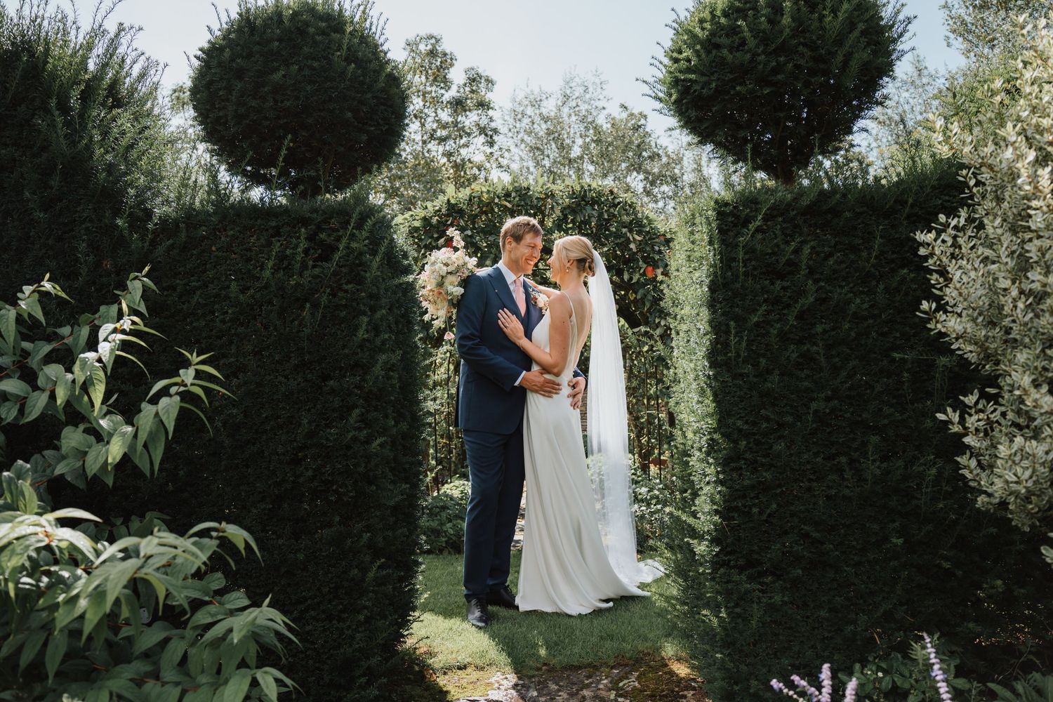 Bride and groom share intimate moment surrounded by tall sculpted hedges in formal garden at Oxleaze Barn.