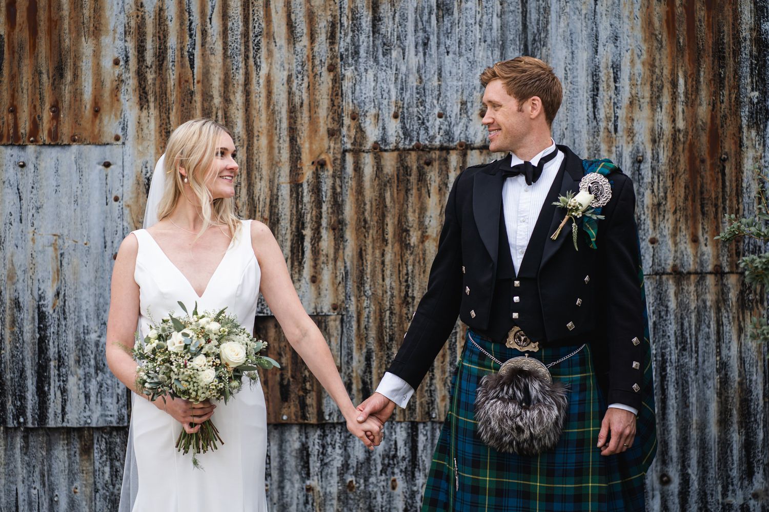 Bride holding bouquet stands with groom wearing a traditional Scottish kilt before a rustic barn Cripps Stone Barn