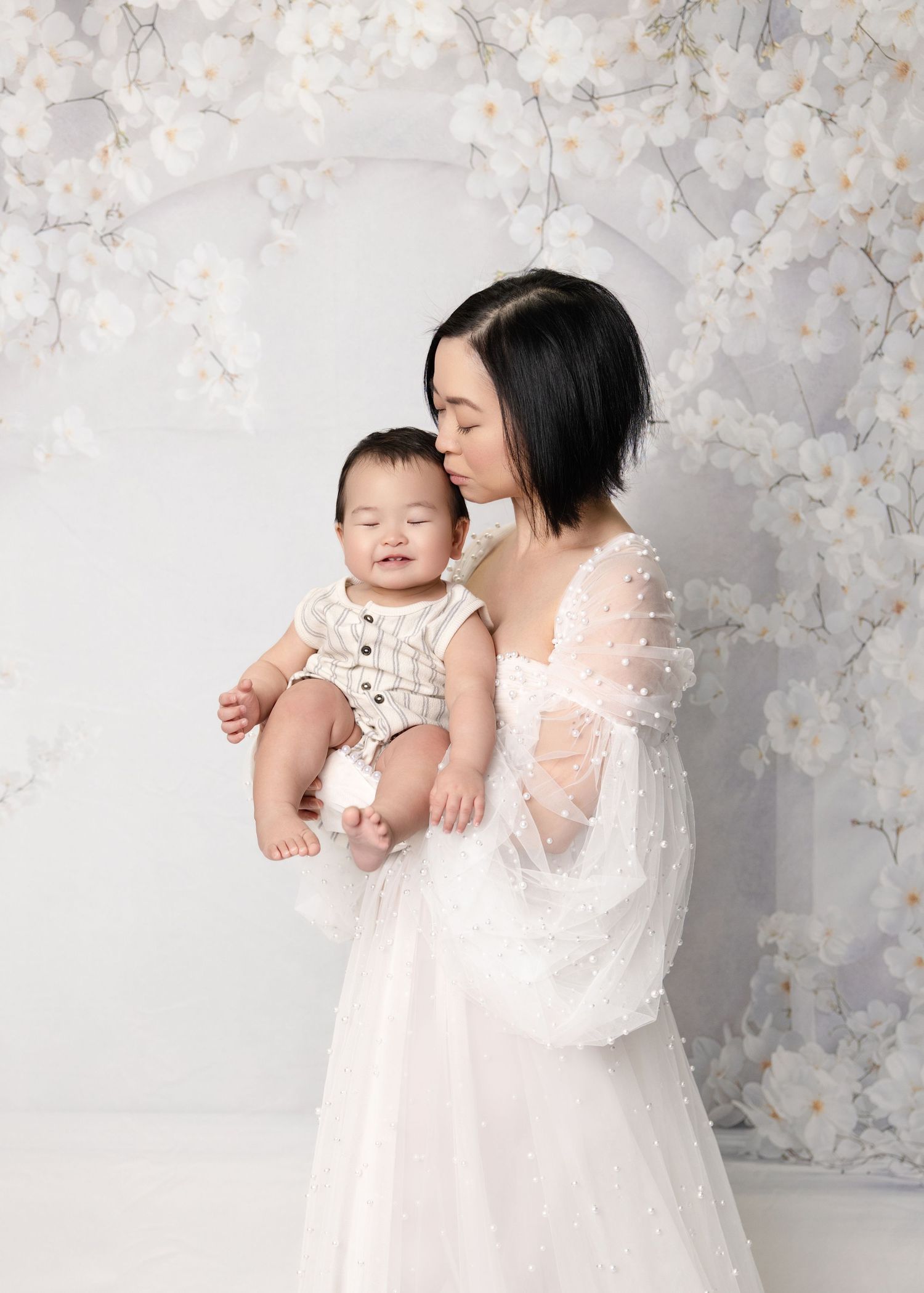 A mother in a white tulle gown kisses her baby dressed in a striped outfit against a floral backdrop.
