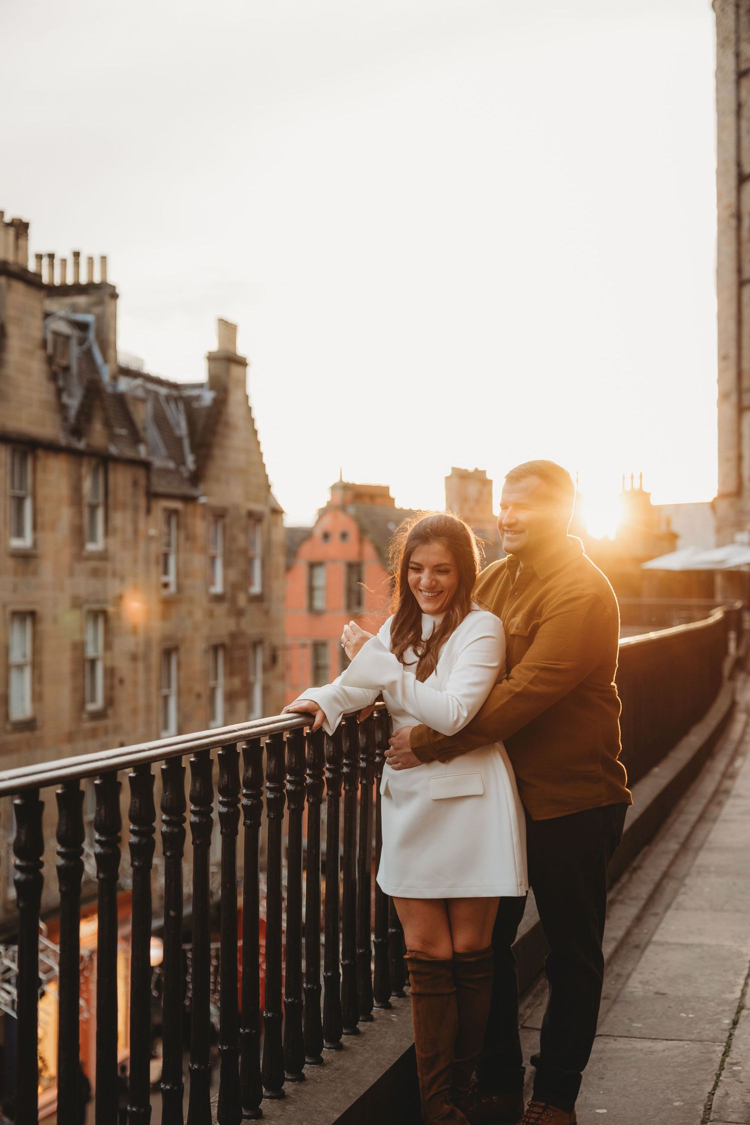Couple embracing on a rooftop balcony at golden sunset with Edinburgh stone buildings in the background.