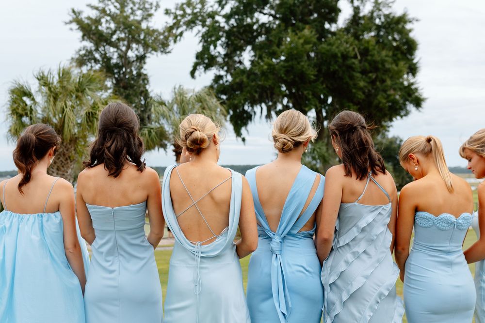 Six bridesmaids in mismatched light blue dresses standing together with backs to camera outdoors.