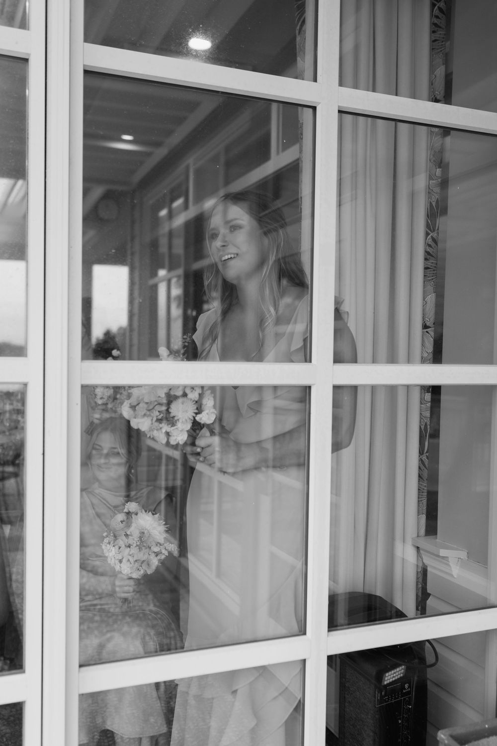 Black and white photo of a smiling bride holding a bouquet, seen through a window with her reflection.