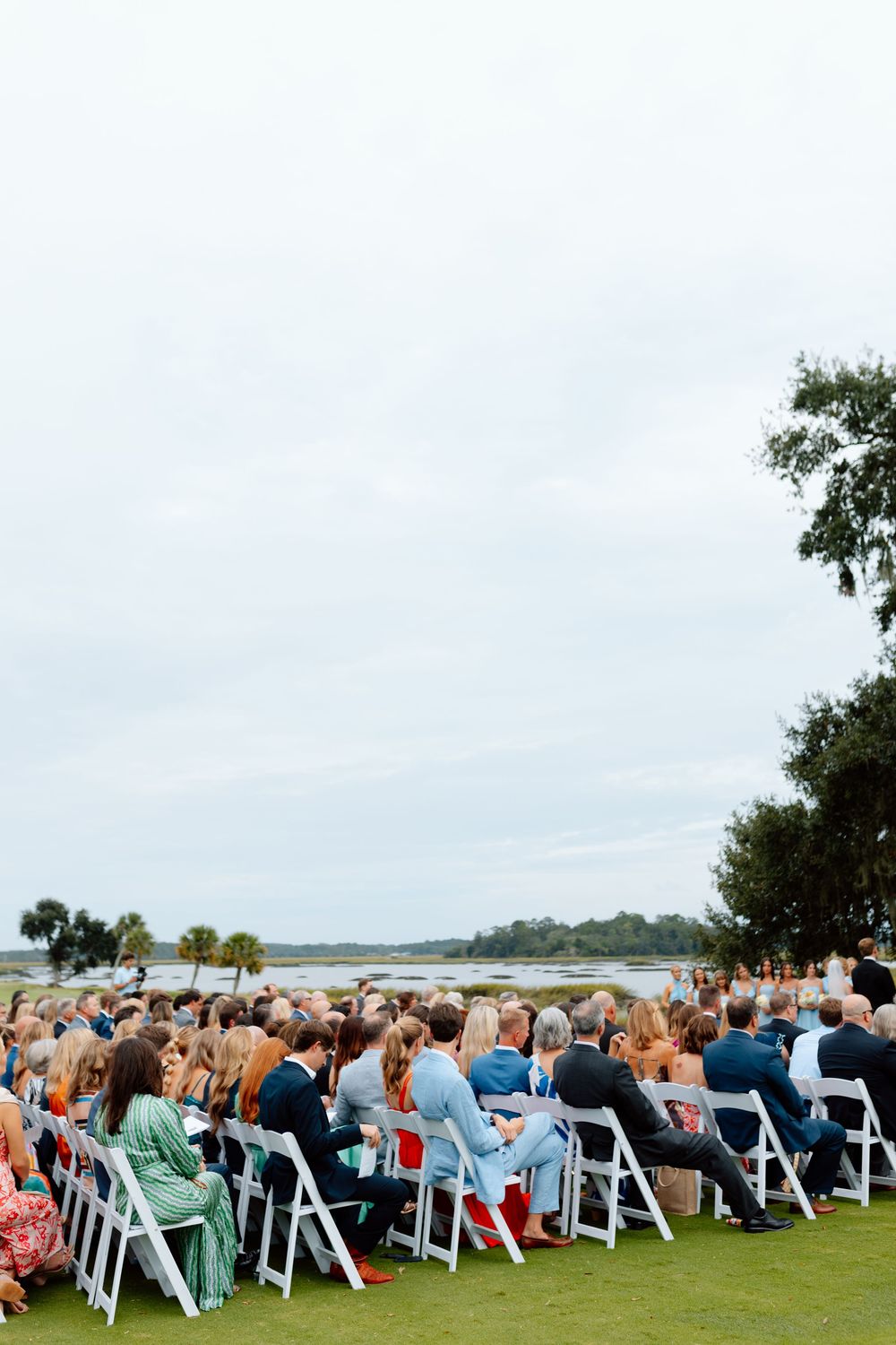 Outdoor wedding ceremony with guests seated in white chairs on a green lawn overlooking a scenic waterway.