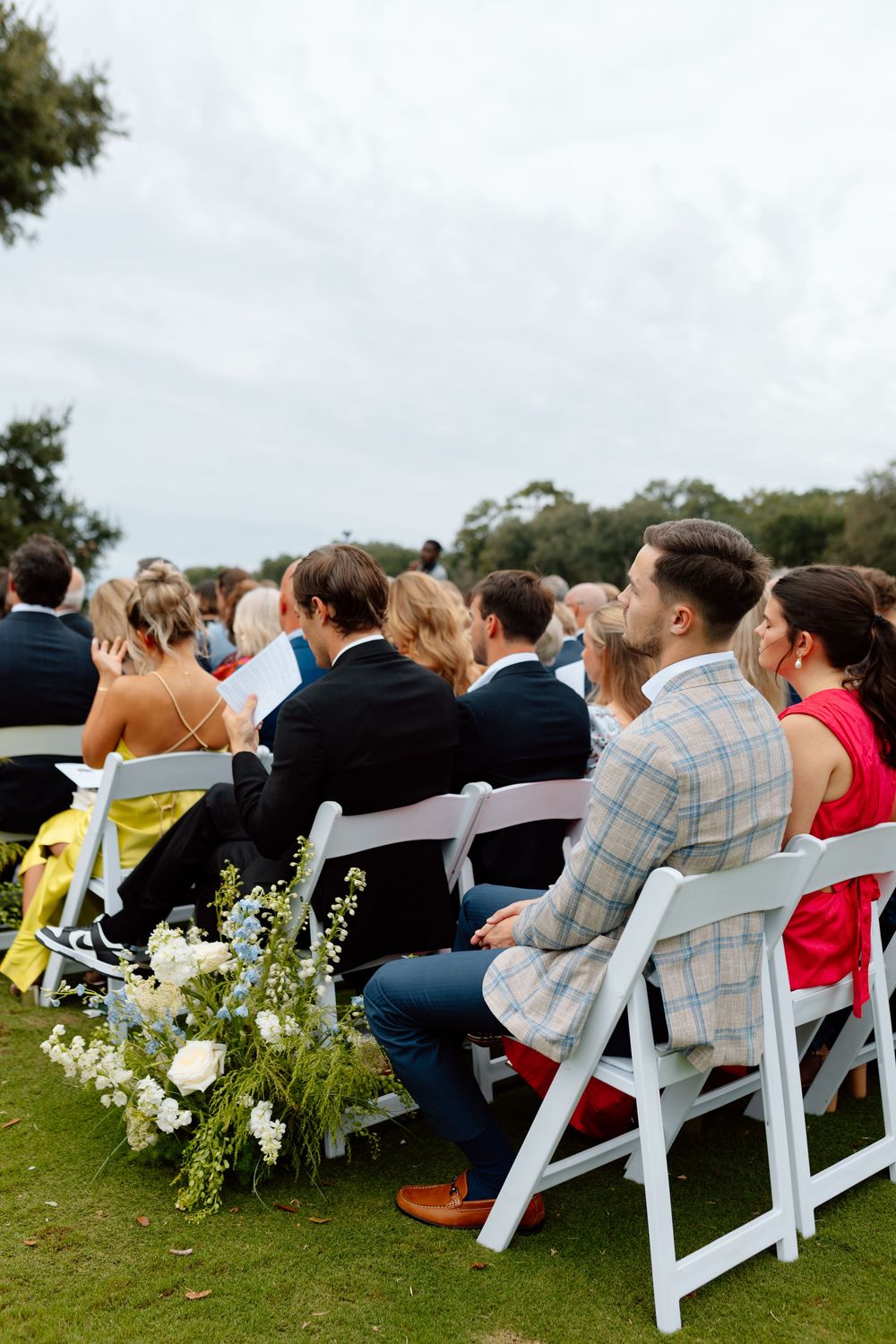 Wedding guests seated in white chairs on a grass lawn, with floral aisle decorations and cloudy sky above.
