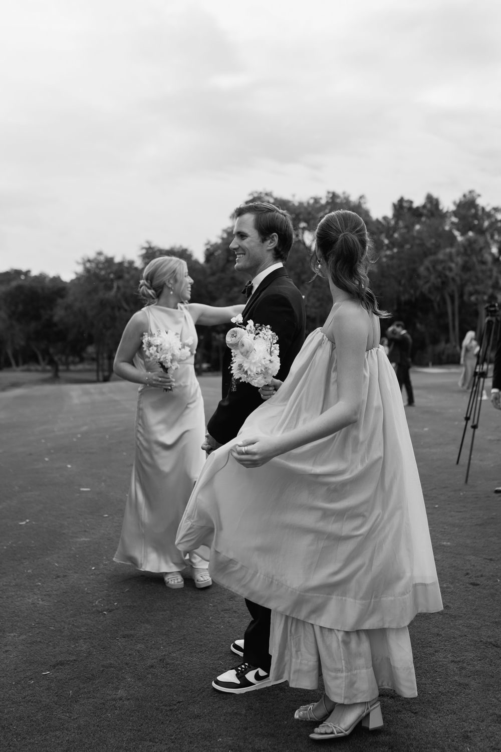 Black and white photo of a groom smiling with two bridesmaids in flowing dresses on an outdoor lawn.