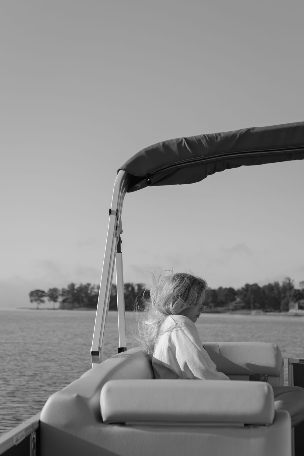 Black and white photo of a child sitting on a pontoon boat on a calm lake with distant trees.