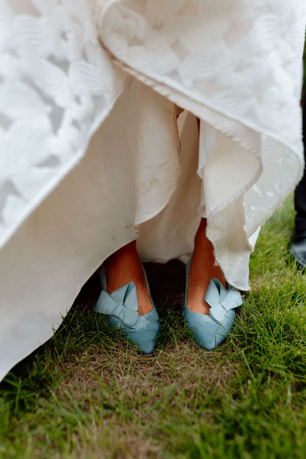 Close-up of a bride wearing light blue bow-detail flats beneath a white lace wedding gown on grass.