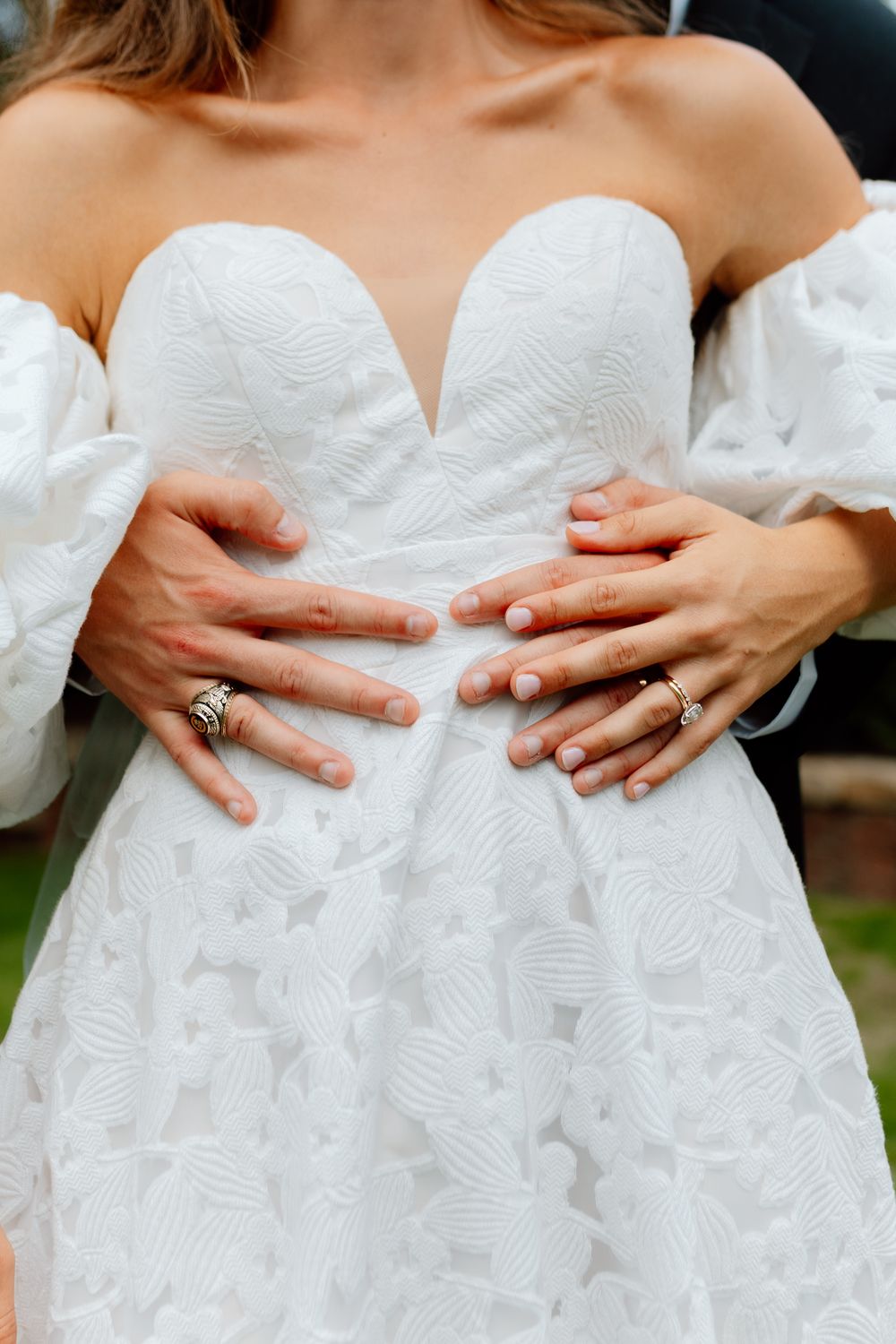 Close-up of a bride and grooms hands with wedding rings resting on her white textured strapless gown.