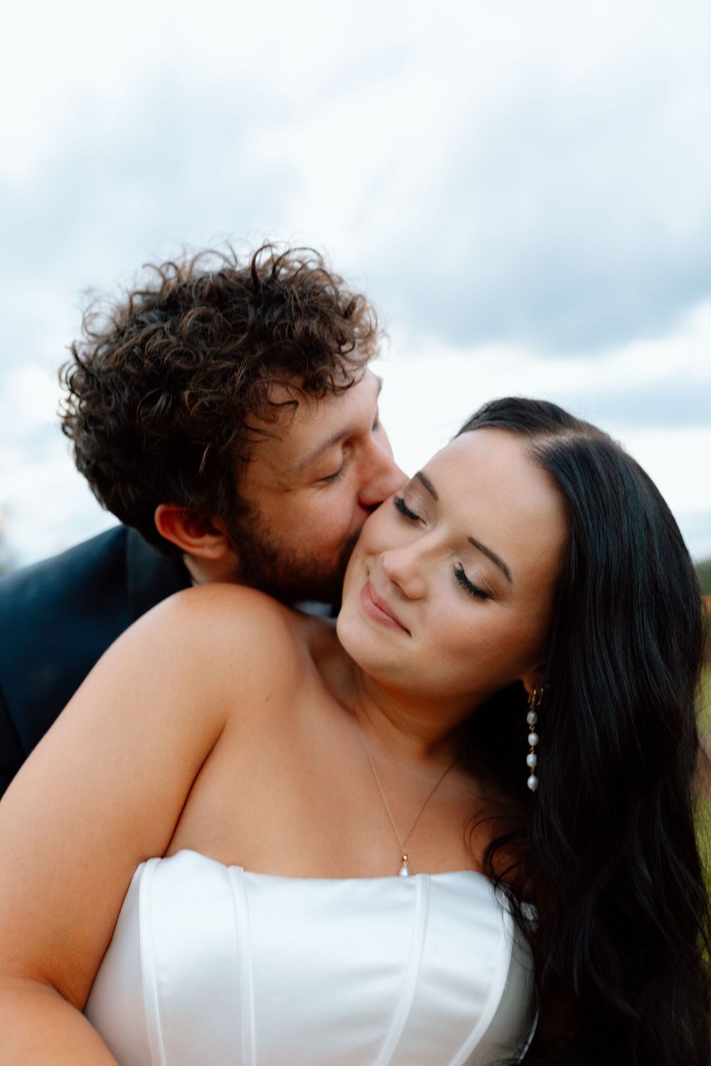 Groom with curly hair tenderly kisses the cheek of his dark-haired bride in a white strapless gown.