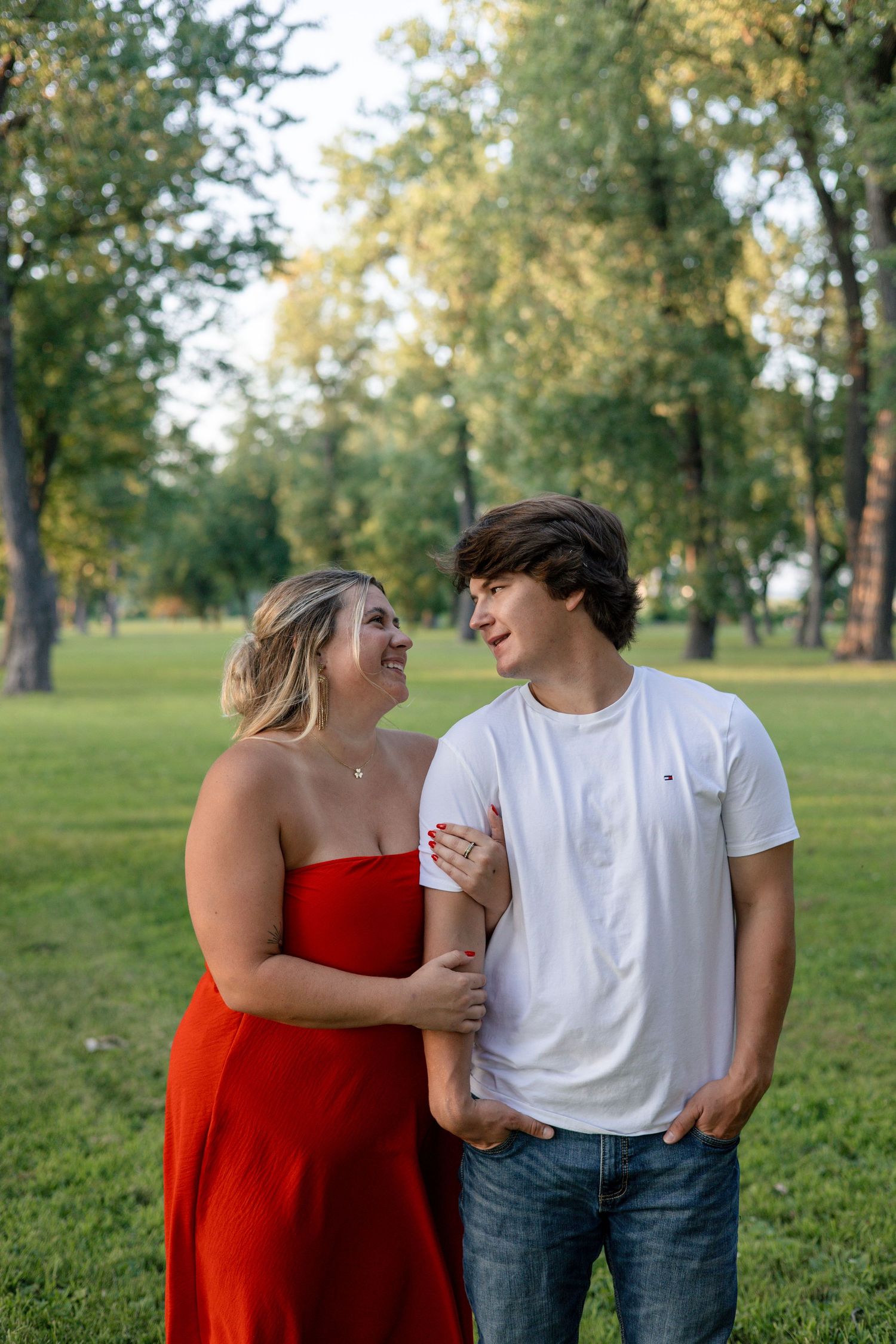 Young couple smiling at each other in a green park, woman in red dress, man in white tee.