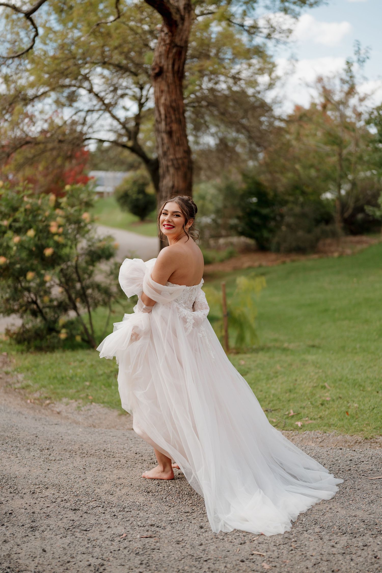 Bride in off-shoulder white tulle gown twirling on gravel path surrounded by lush green trees and colorful foliage.