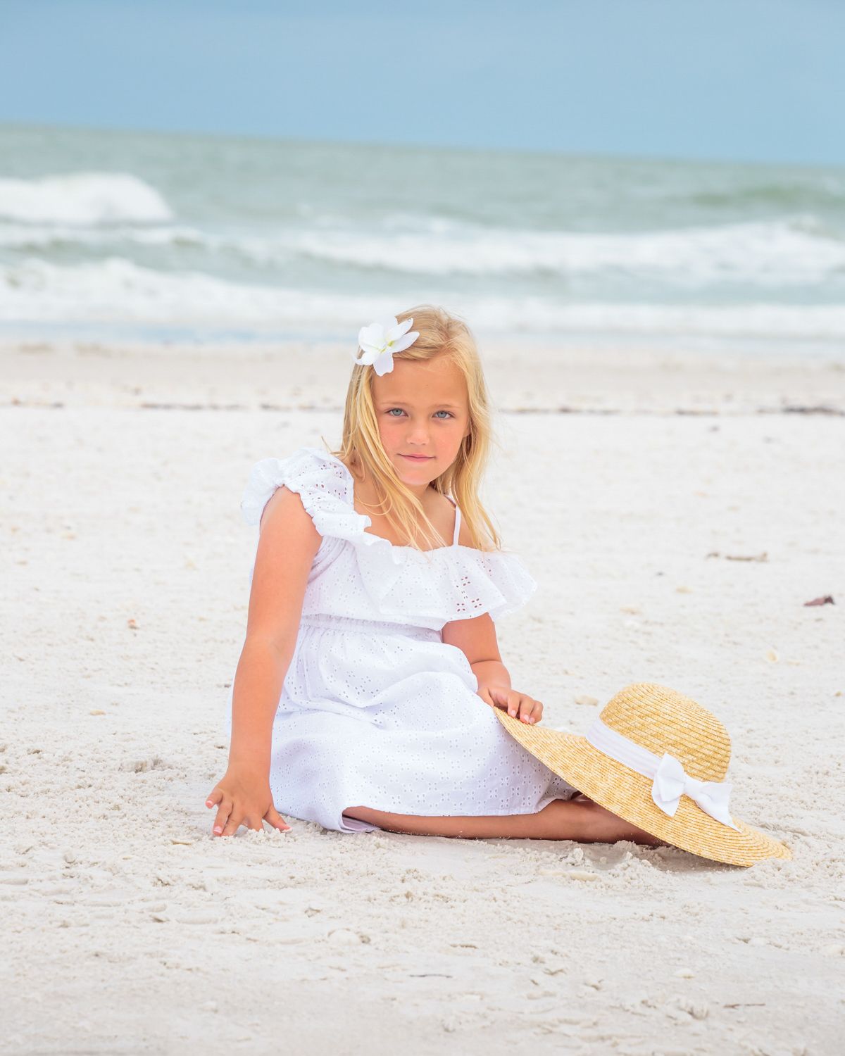 Young girl in white dress reclining on sandy beach in Naples FL with sun hat and ocean waves in background, by Mark Block Photography