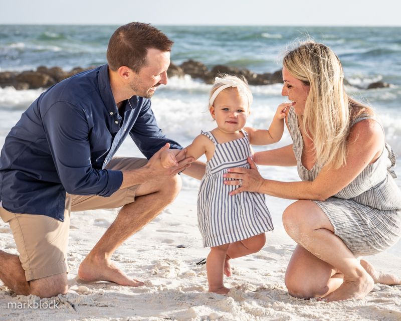 Candid family photography on a sunny Naples FL beach as parents help their toddler in a striped dress take first steps.