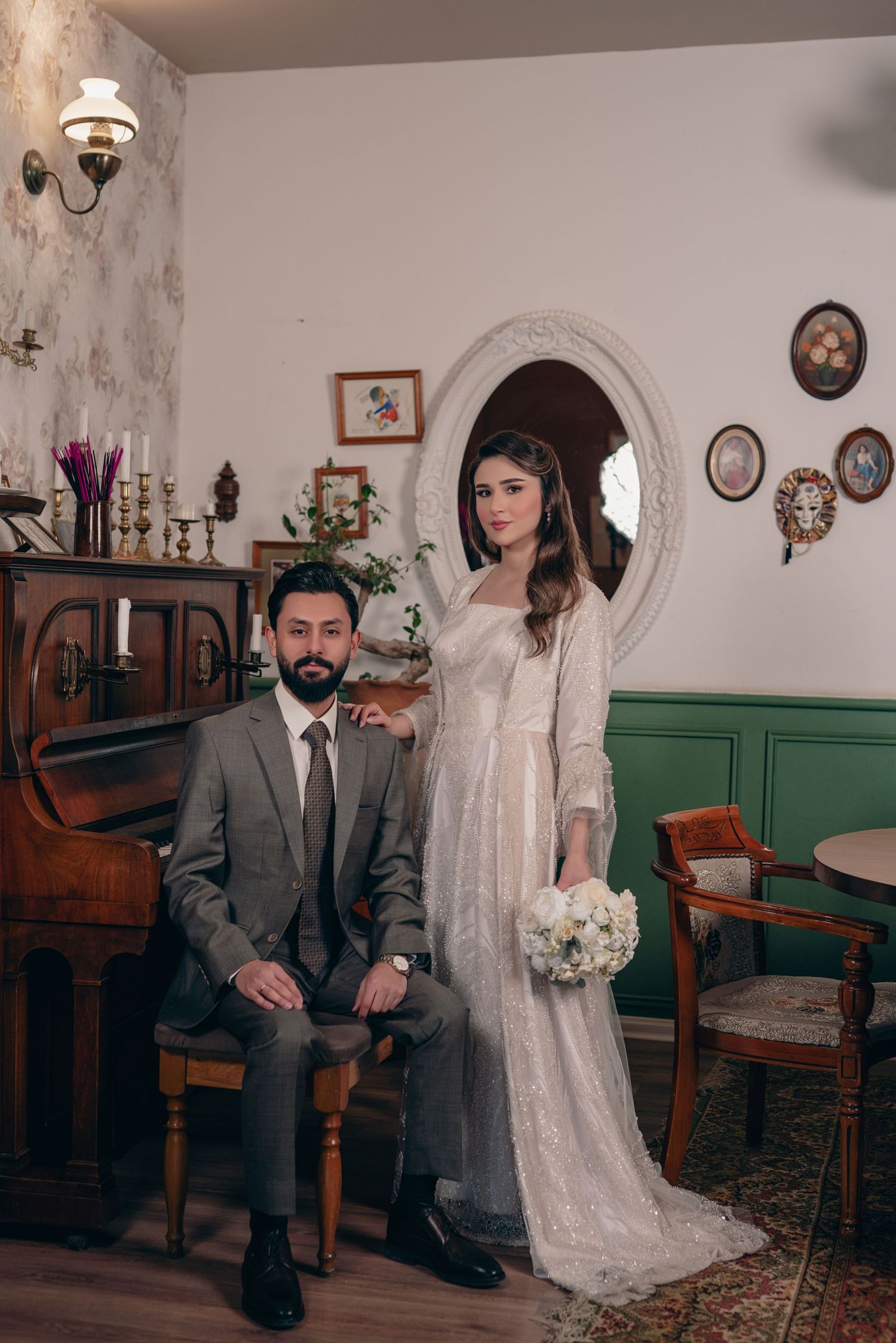 A couple in vintage-inspired wedding attire pose in an antique-decorated room with ornate furniture and wall decor.