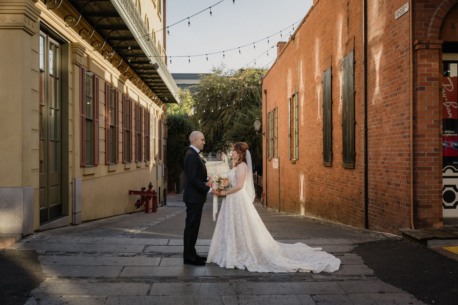 Wedding couple stands in a rustic brick alleyway decorated with string lights and industrial charm for Sacramento photo session