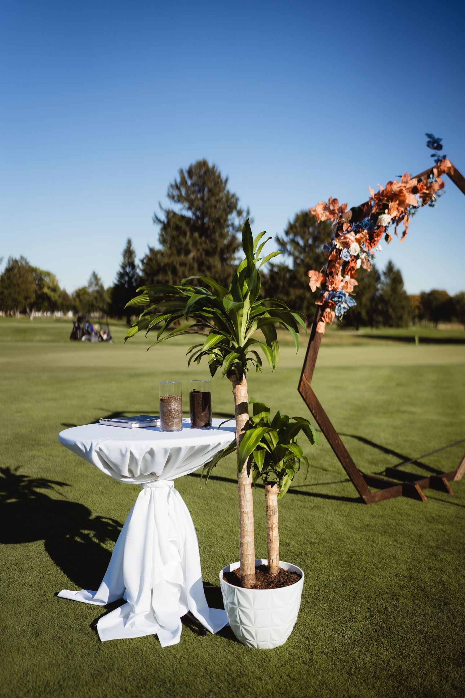 A cocktail table with white linens and a potted plant sits on a golf course with trees in the background.