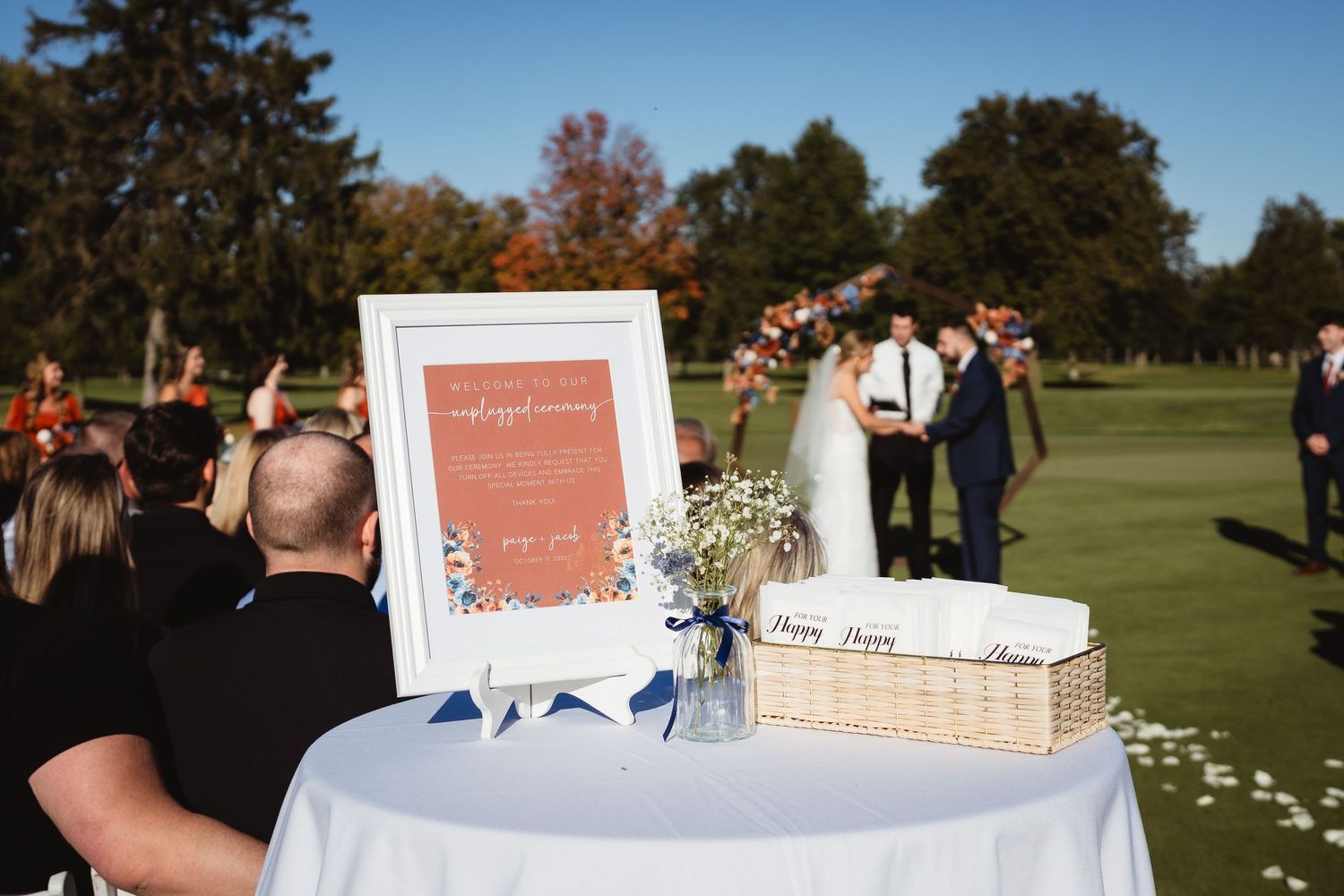 Outdoor wedding ceremony setup with white welcome sign and decorative elements on round table at golf course.