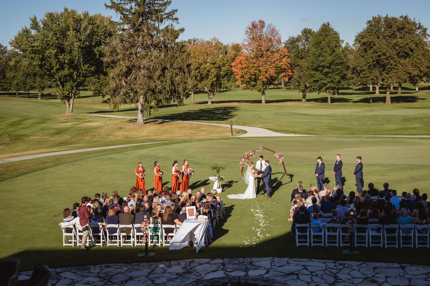 An outdoor wedding ceremony setup on a golf course with rows of white chairs and autumn trees in the background.