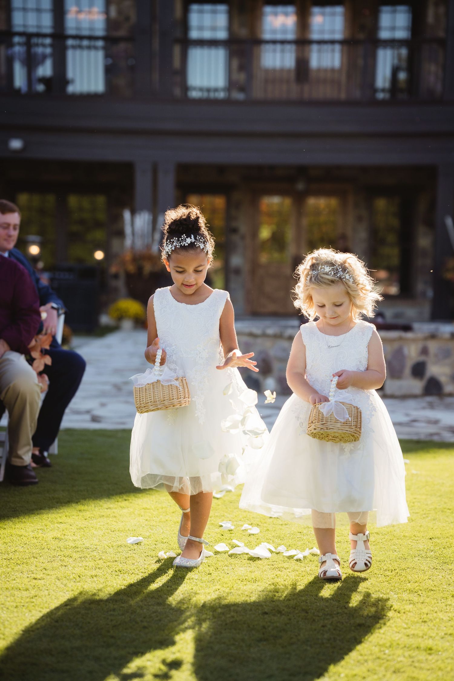 Young flower girls in white dresses scatter petals on green grass during an outdoor wedding ceremony.