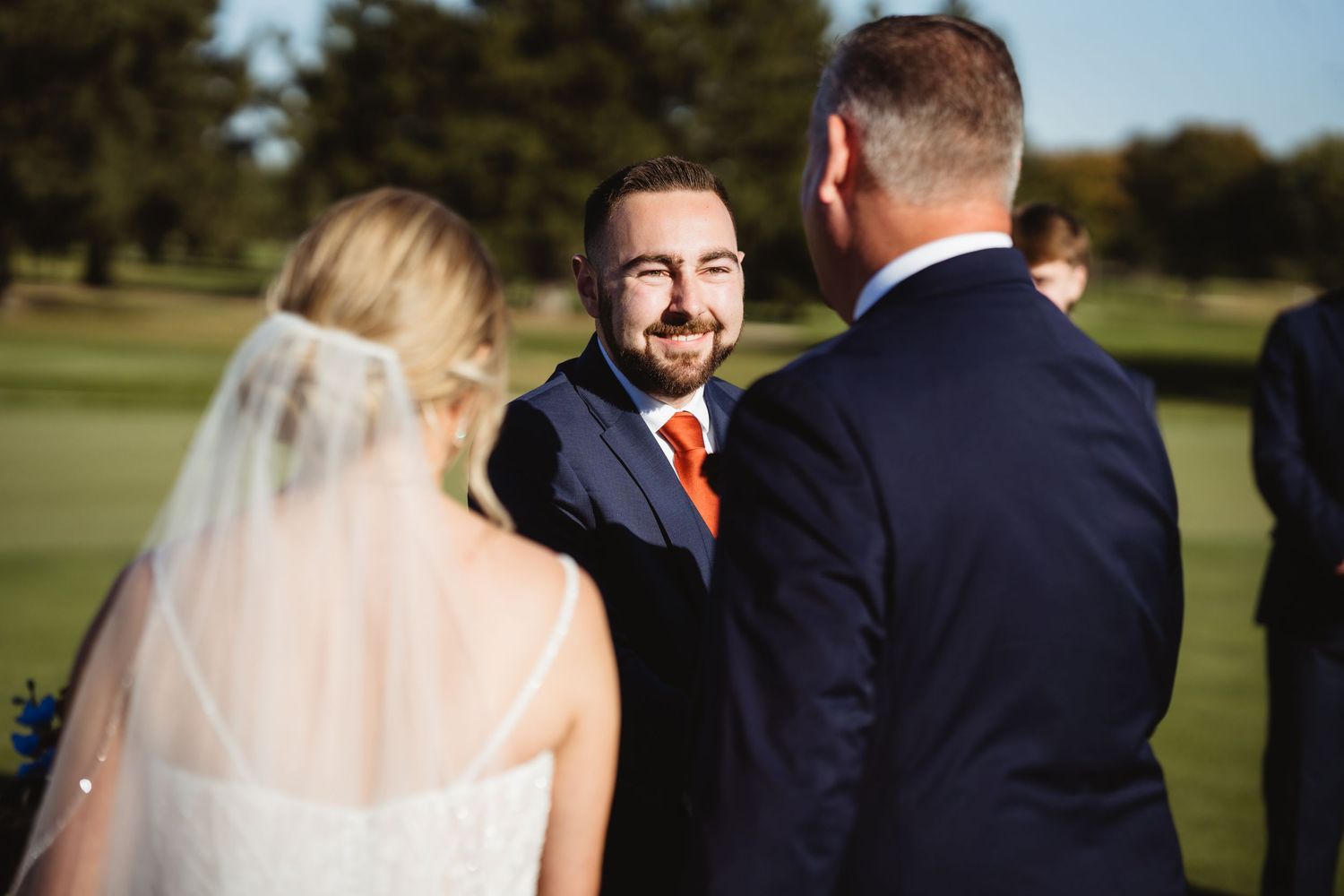 A wedding party poses together on a sunny golf course with trees in the background.