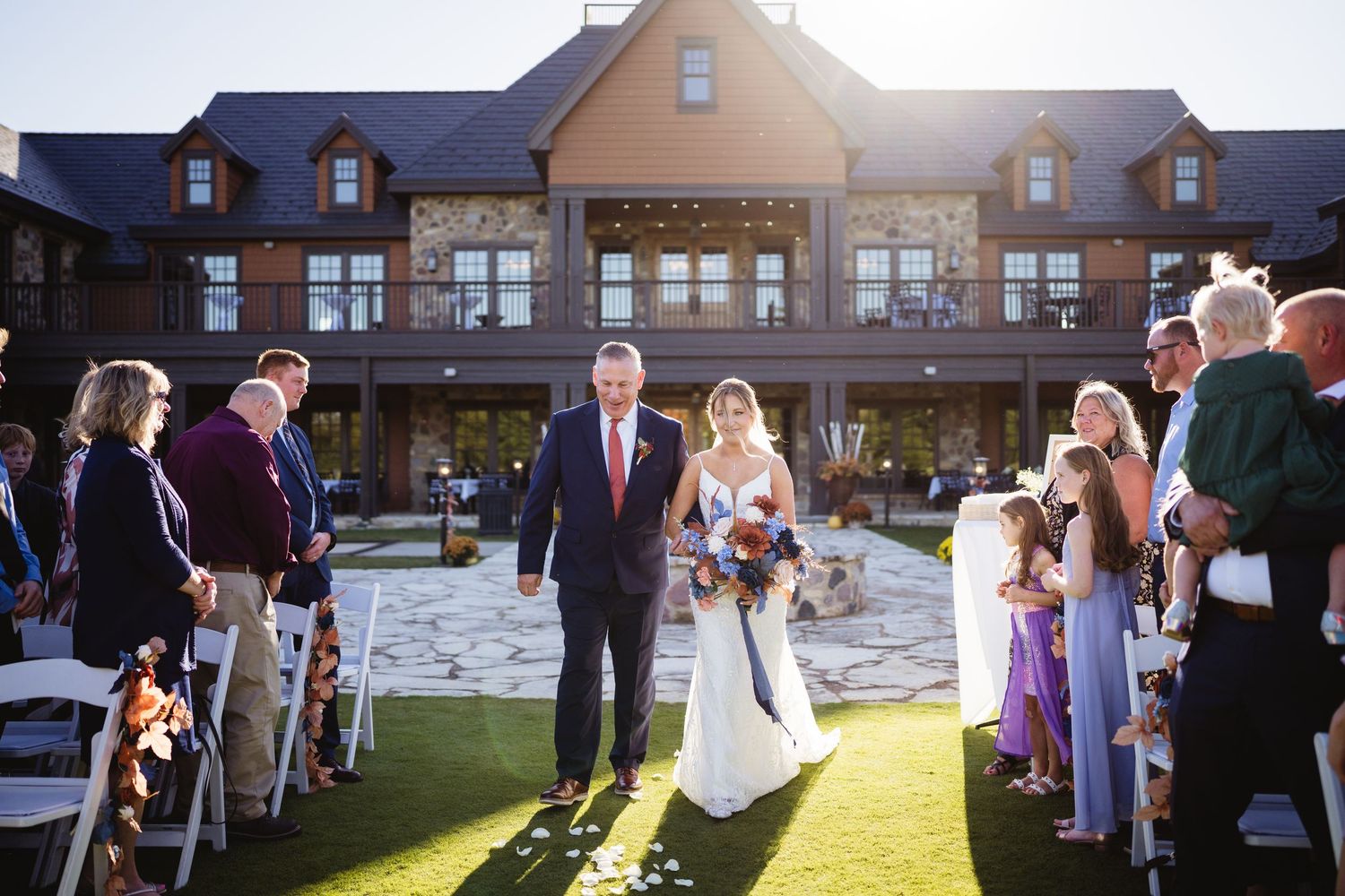 A wedding ceremony takes place on a sunny lawn in front of a grand lodge-style building.