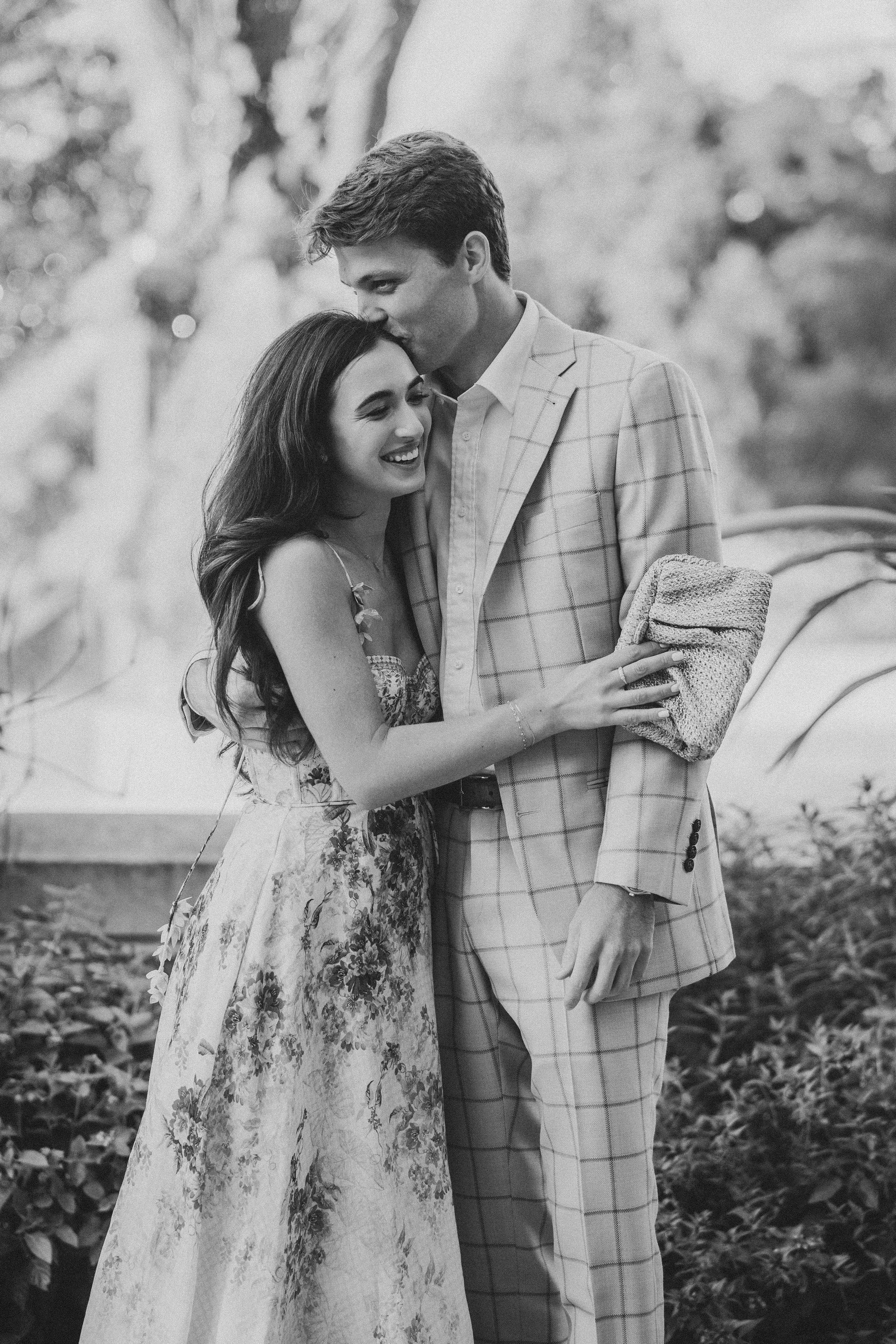 A romantic couple in a garden shares intimate moments wearing a floral dress and plaid blazer in black and white photography.