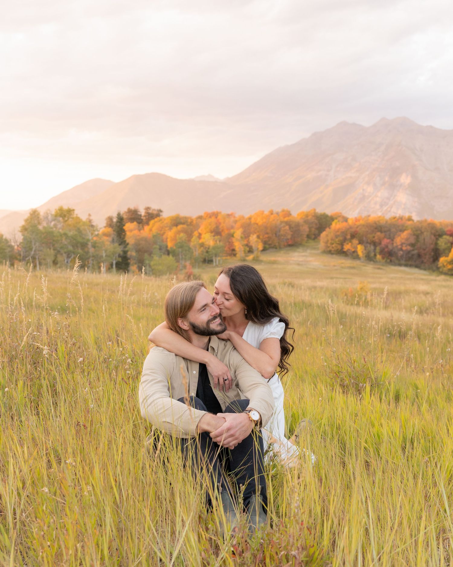 A couple embraces lovingly in a golden autumn field at sunset with mountains and fall foliage in the background.