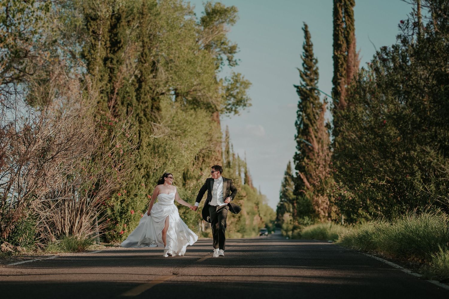 A couple in wedding attire walks together along a tree-lined path during a golden autumn day.