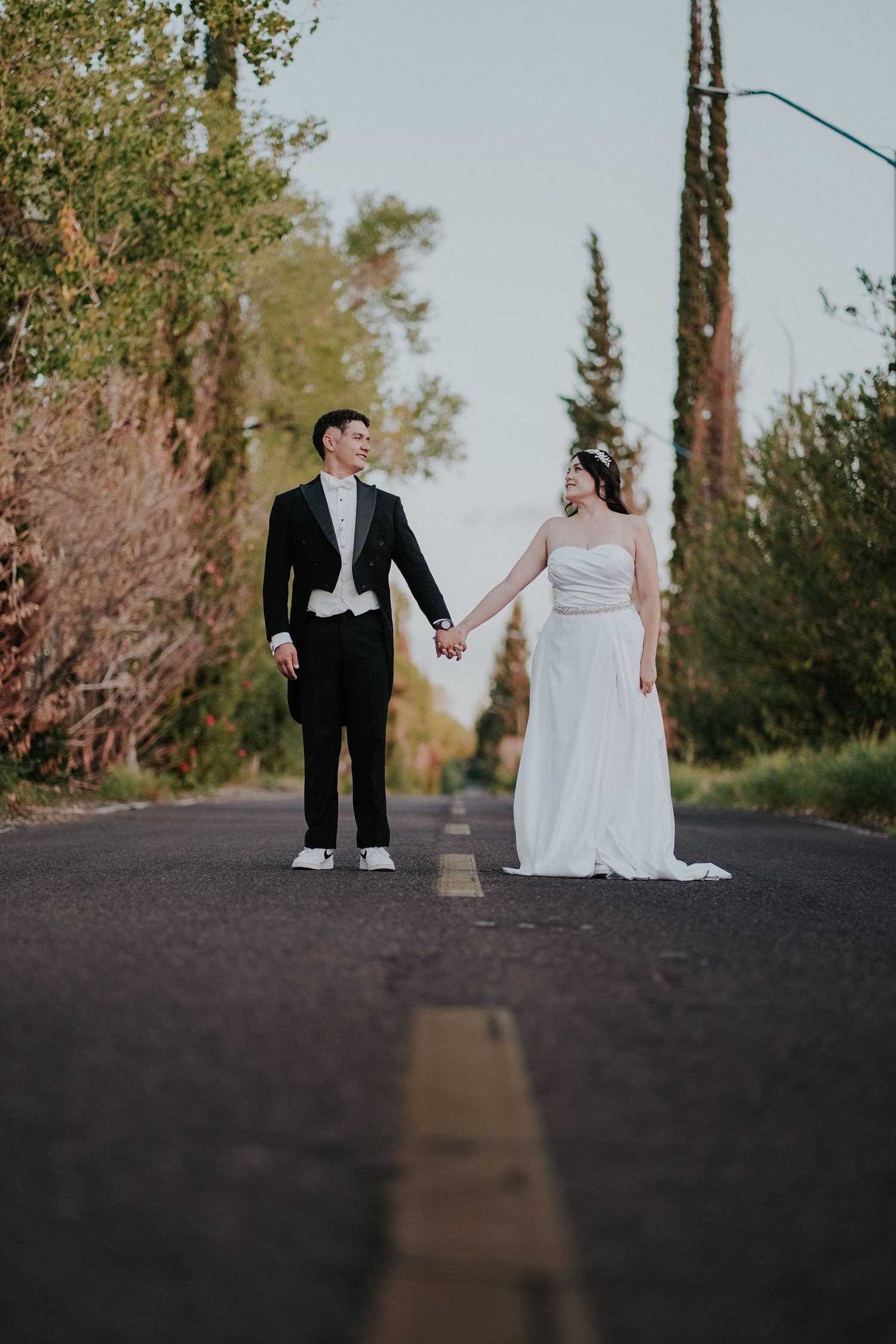 A bride and groom share a playful moment sitting on pavement surrounded by tall trees in an outdoor setting.