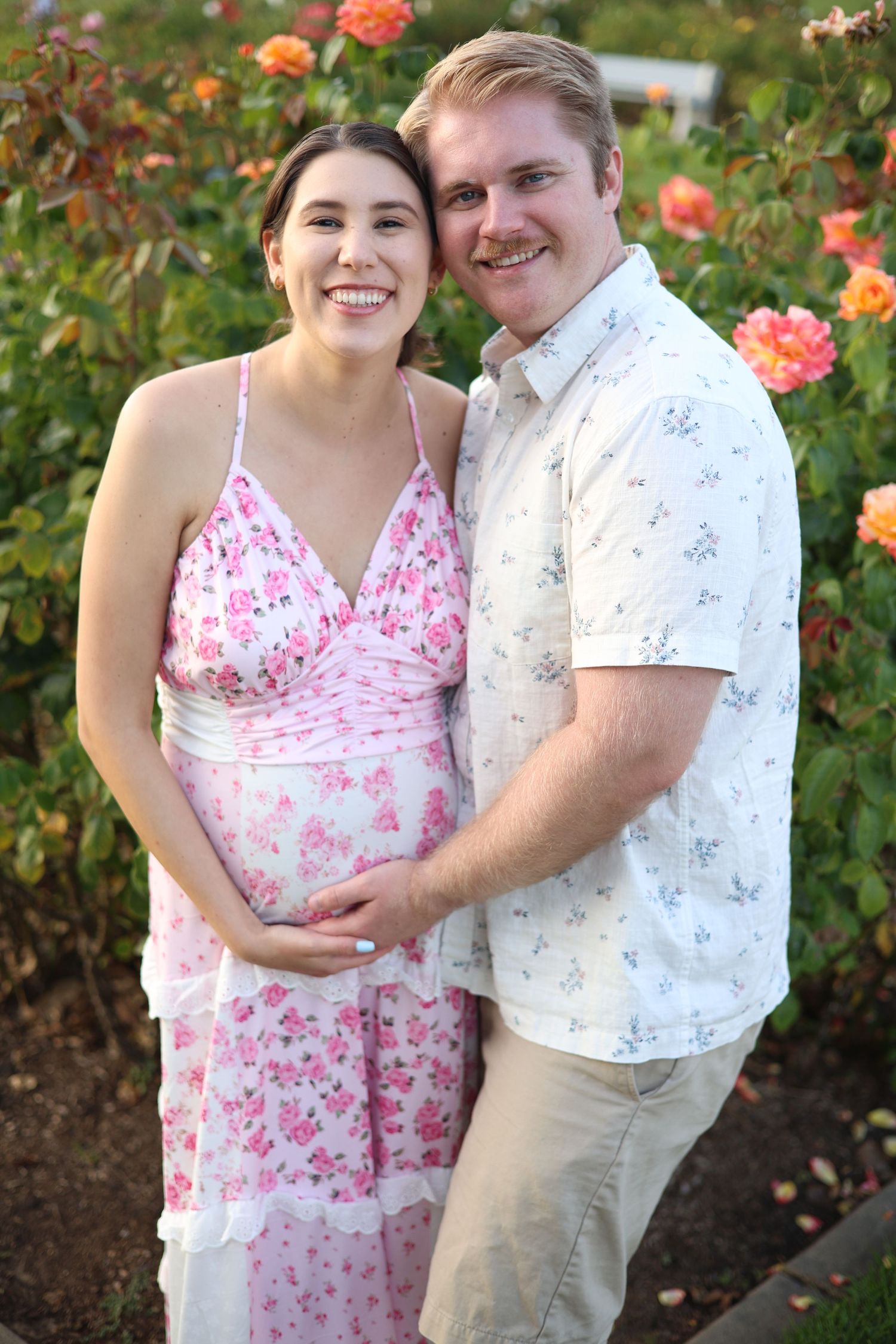 Smiling pregnant woman in pink floral dress embraced by man in floral shirt standing in front of rose bushes.