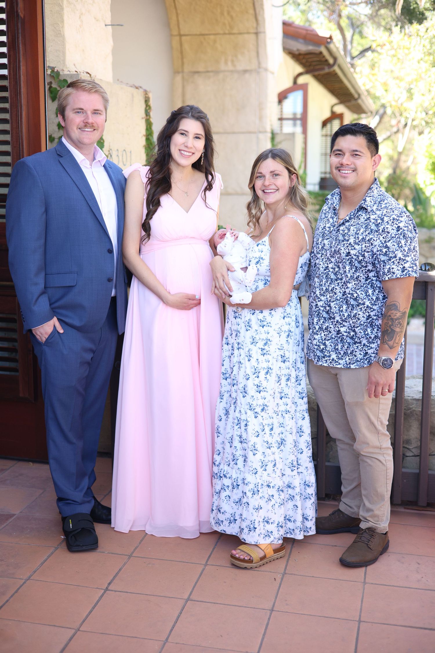 Group of four adults posing outdoors, including a pregnant woman in pink gown and woman holding a newborn baby.
