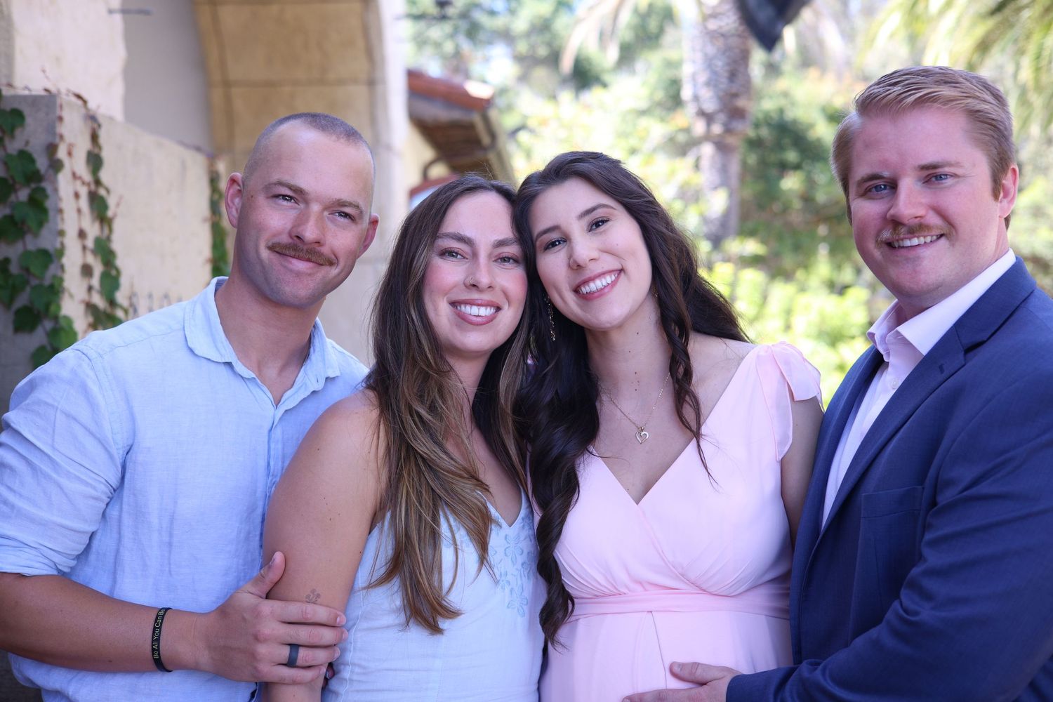 Two smiling couples pose closely together outdoors with palm trees and greenery in the background.