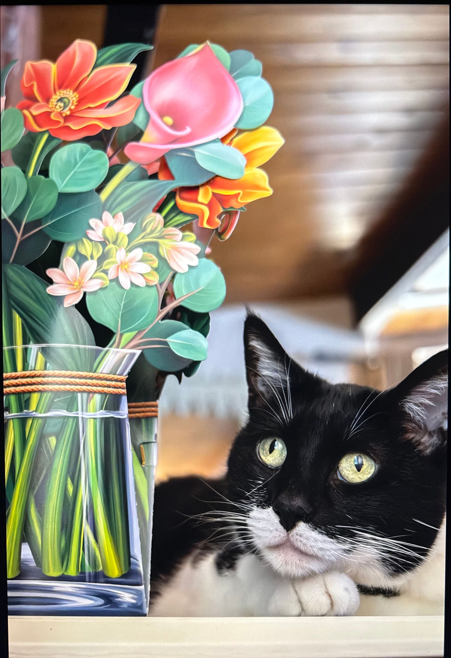Black and white tuxedo cat rests beside a glass vase of colorful painted flowers on a wooden surface.