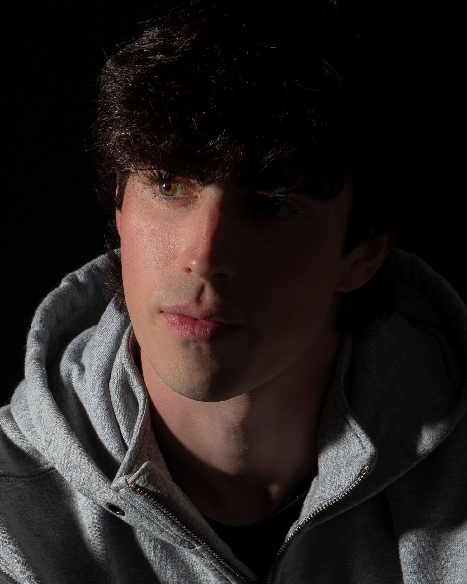 Dramatic low-key portrait of young man with dark hair wearing gray hoodie against black background.