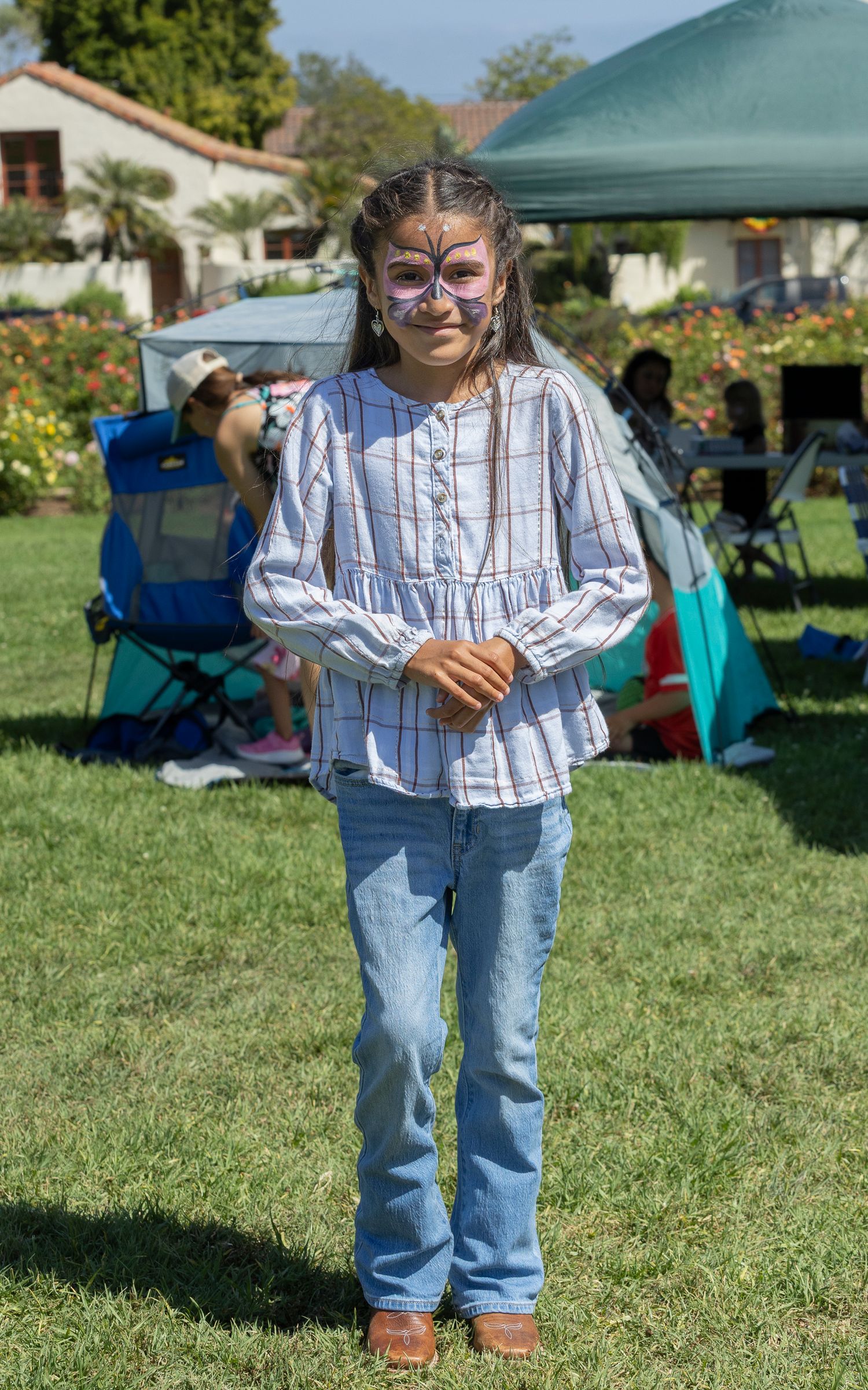 A young girl with butterfly face paint stands on grass at an outdoor event, wearing a plaid top and jeans.