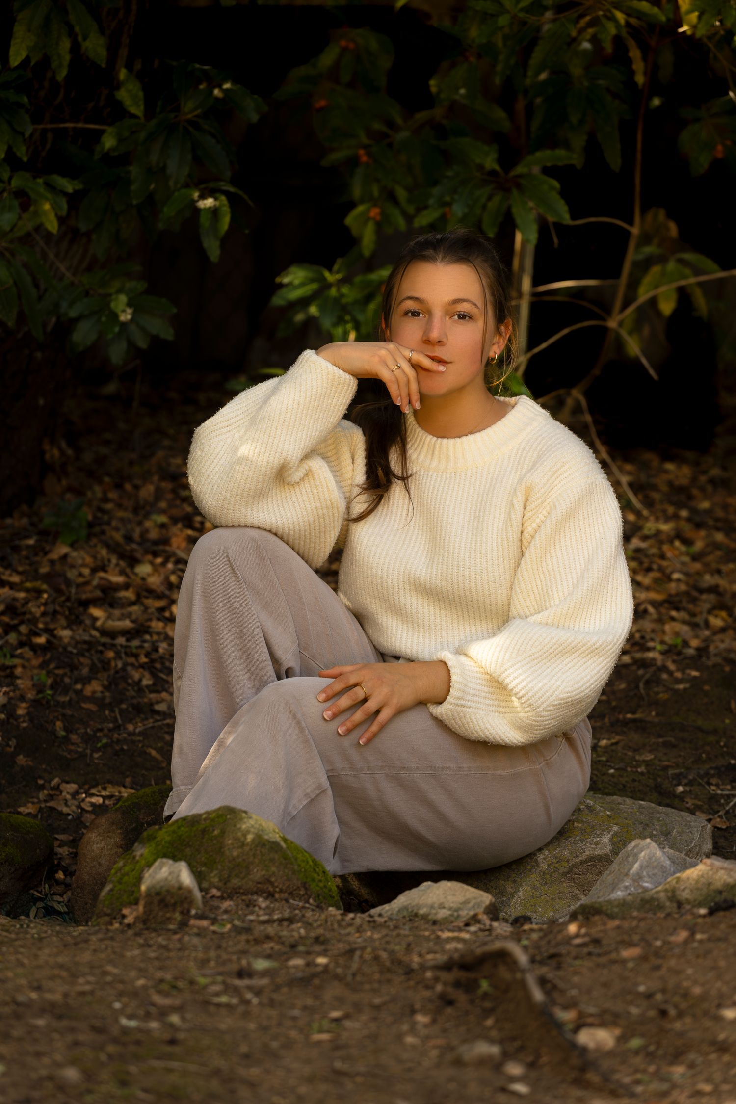 Young woman in cream knit sweater and gray pants sits on rocks outdoors in warm golden sunlight.