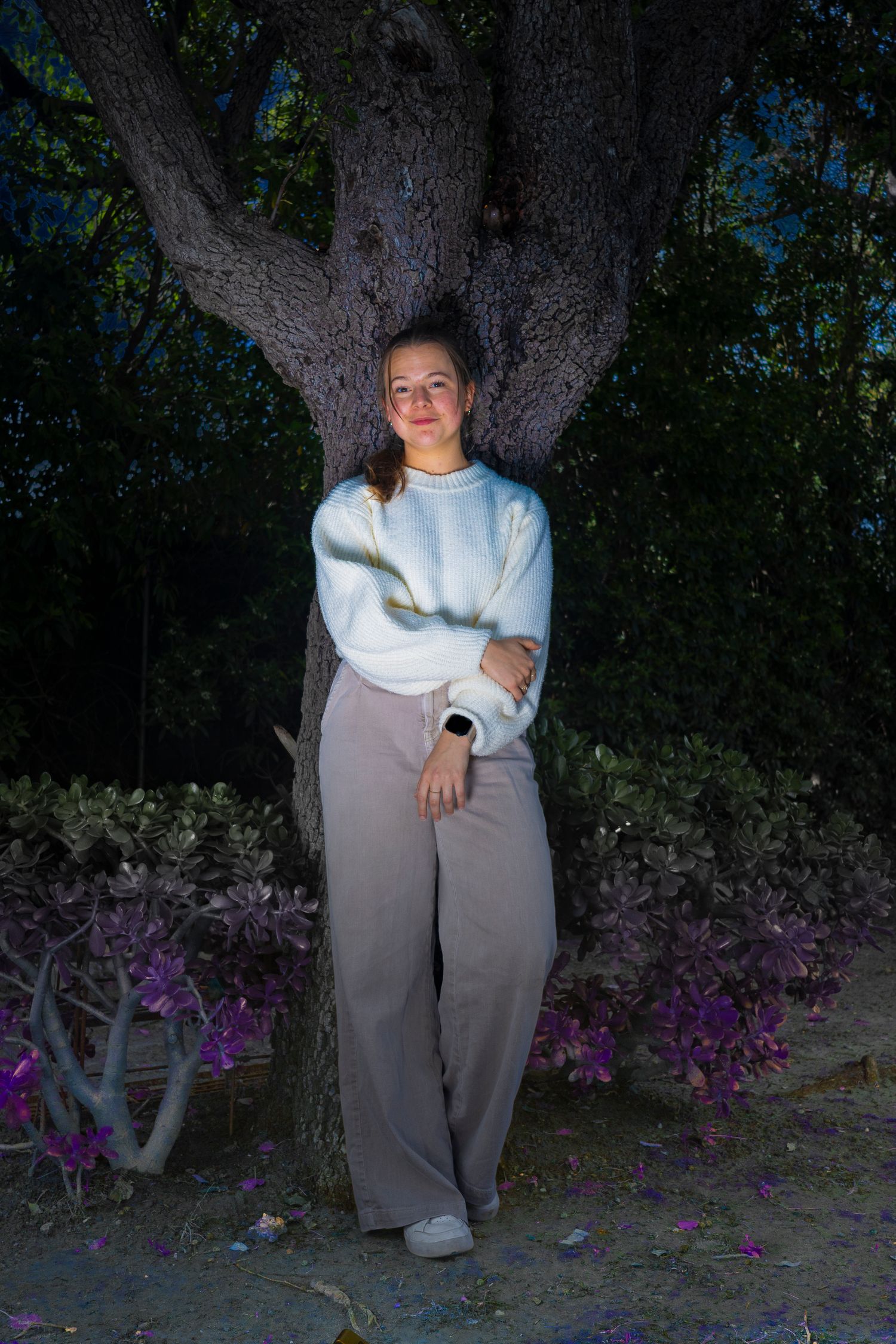 Young woman in white sweater and gray wide-leg pants leans against a large tree at night surrounded by purple flowers.