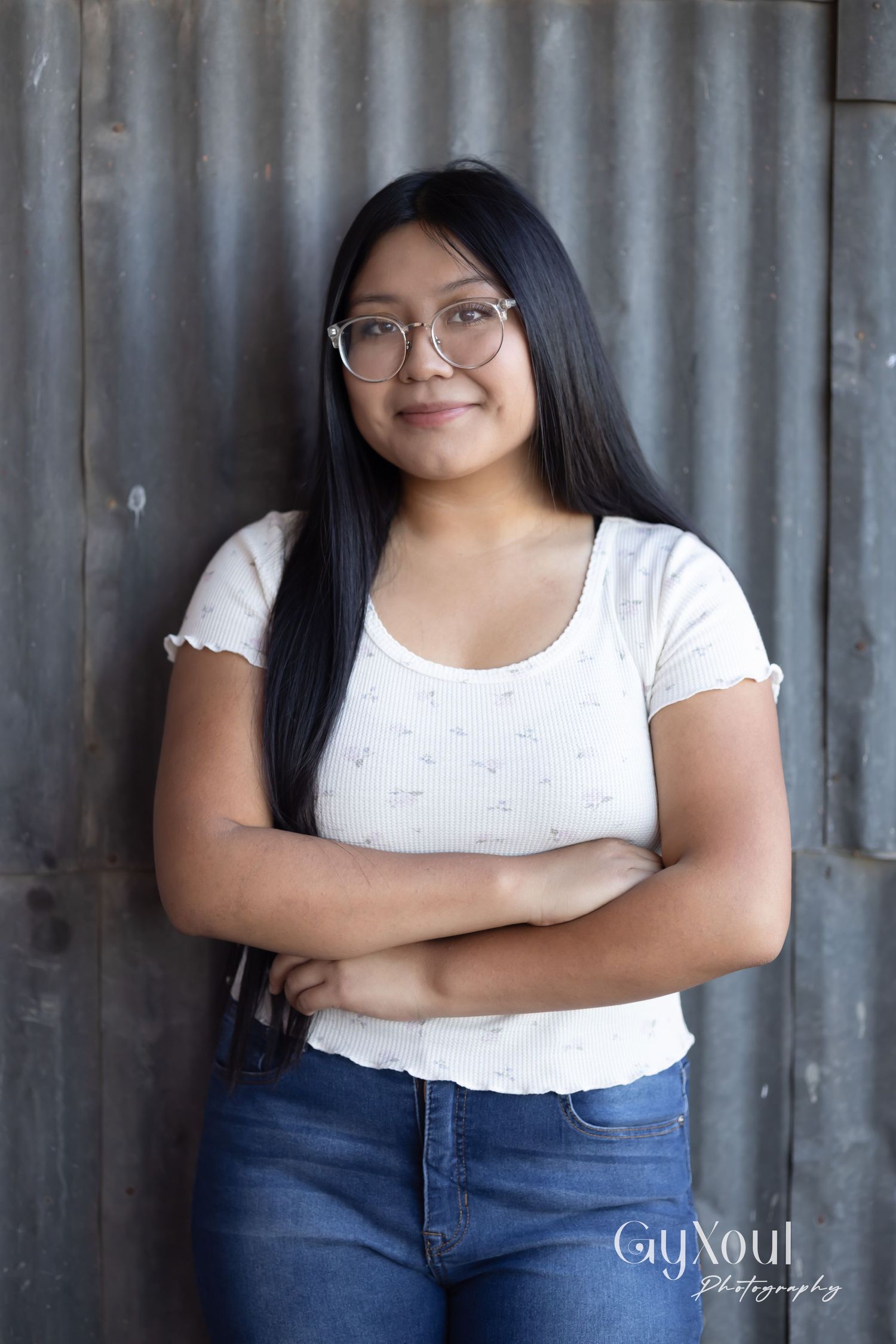 Portrait against metal wall showing person in white top and jeans with crossed arms.