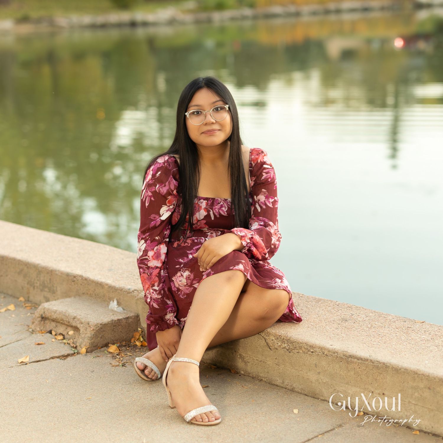 Person in floral burgundy dress sitting by lake on concrete ledge.
