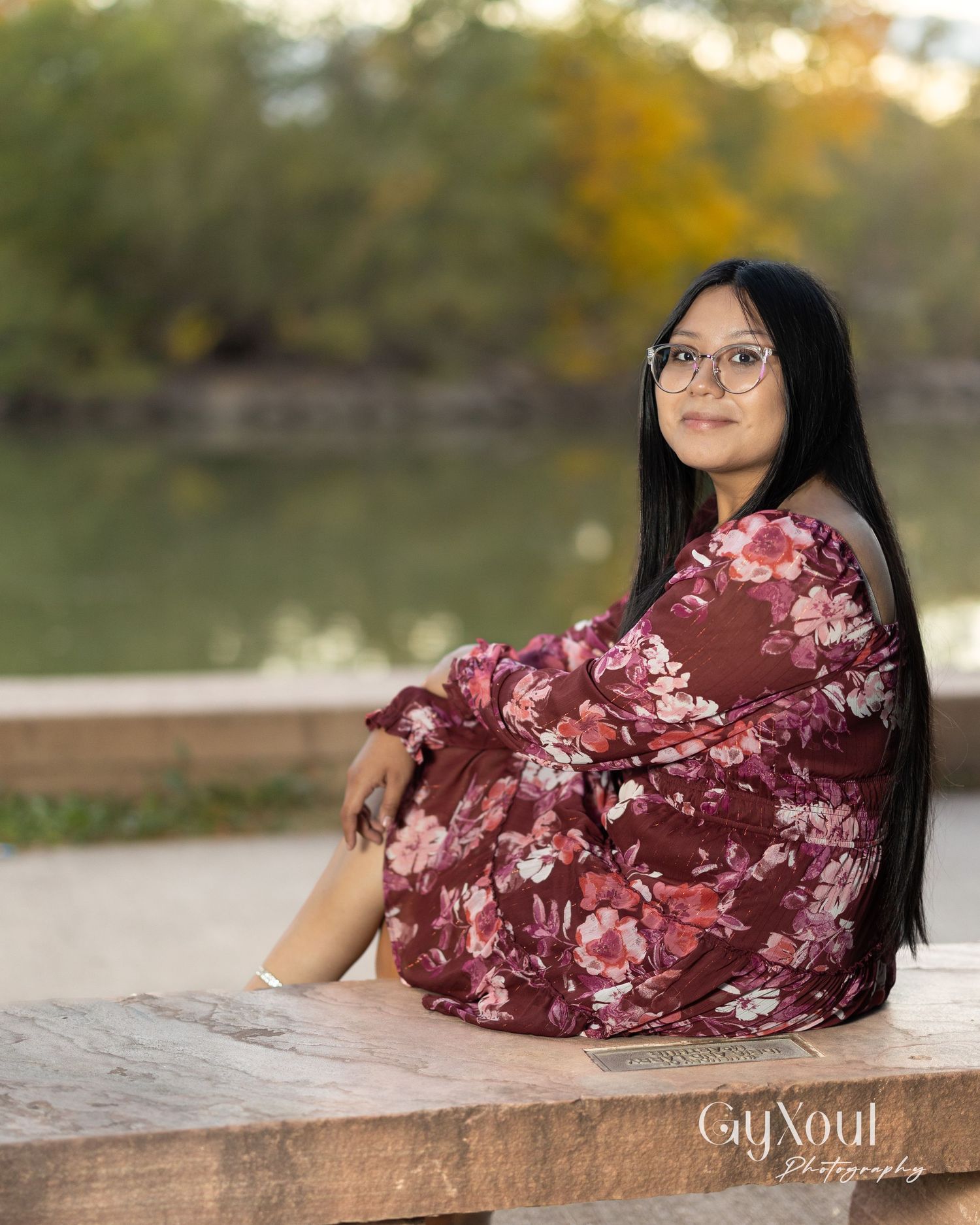 Person in floral burgundy dress sitting by pond with autumn foliage in background.