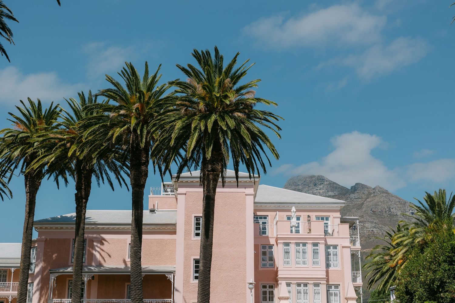 Pink stucco buildings with palm trees line a street against a backdrop of mountains and blue sky.