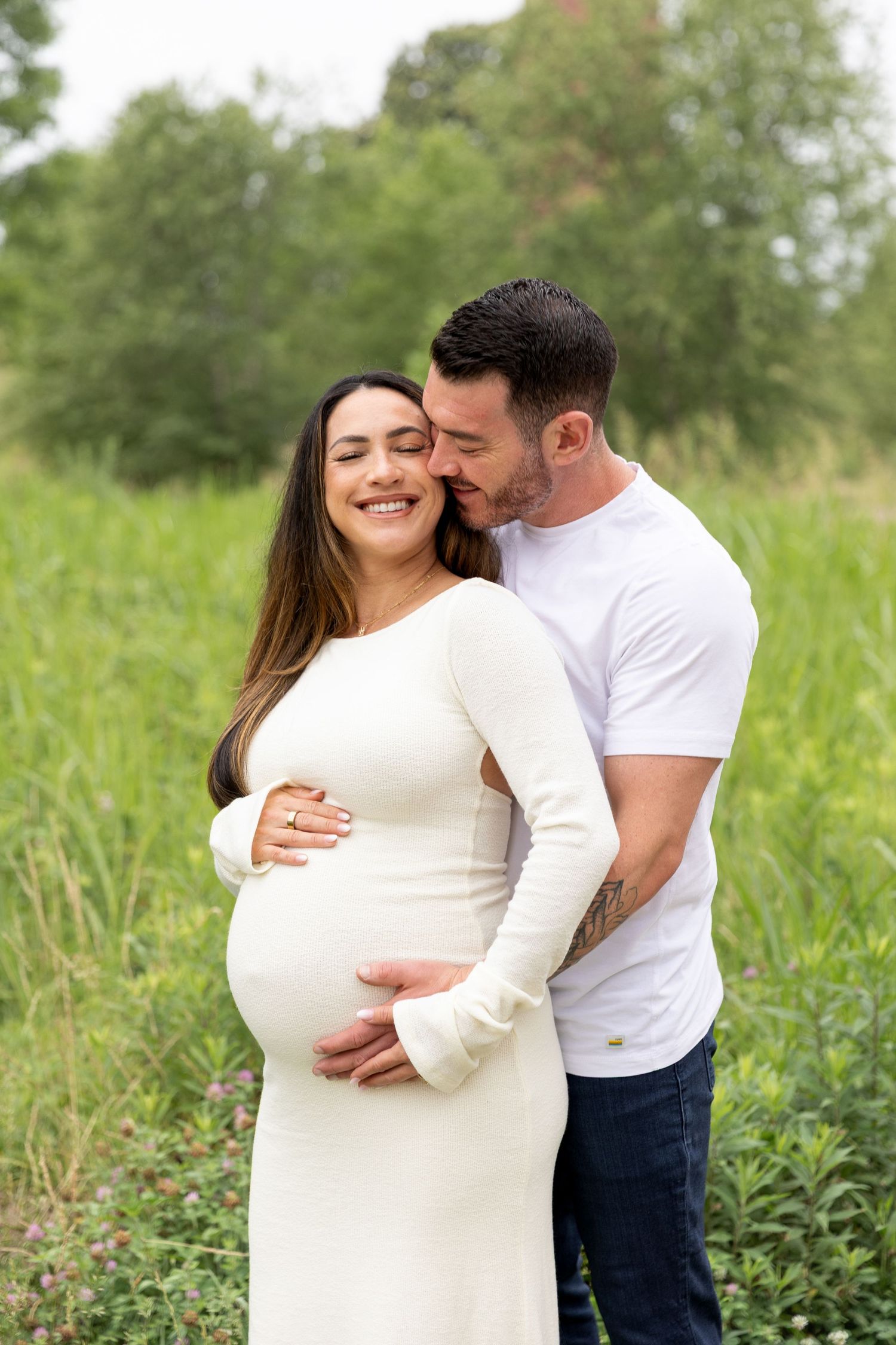 Couple embracing outdoors, pregnant woman in white dress smiling, partner kissing her temple in lush green field.