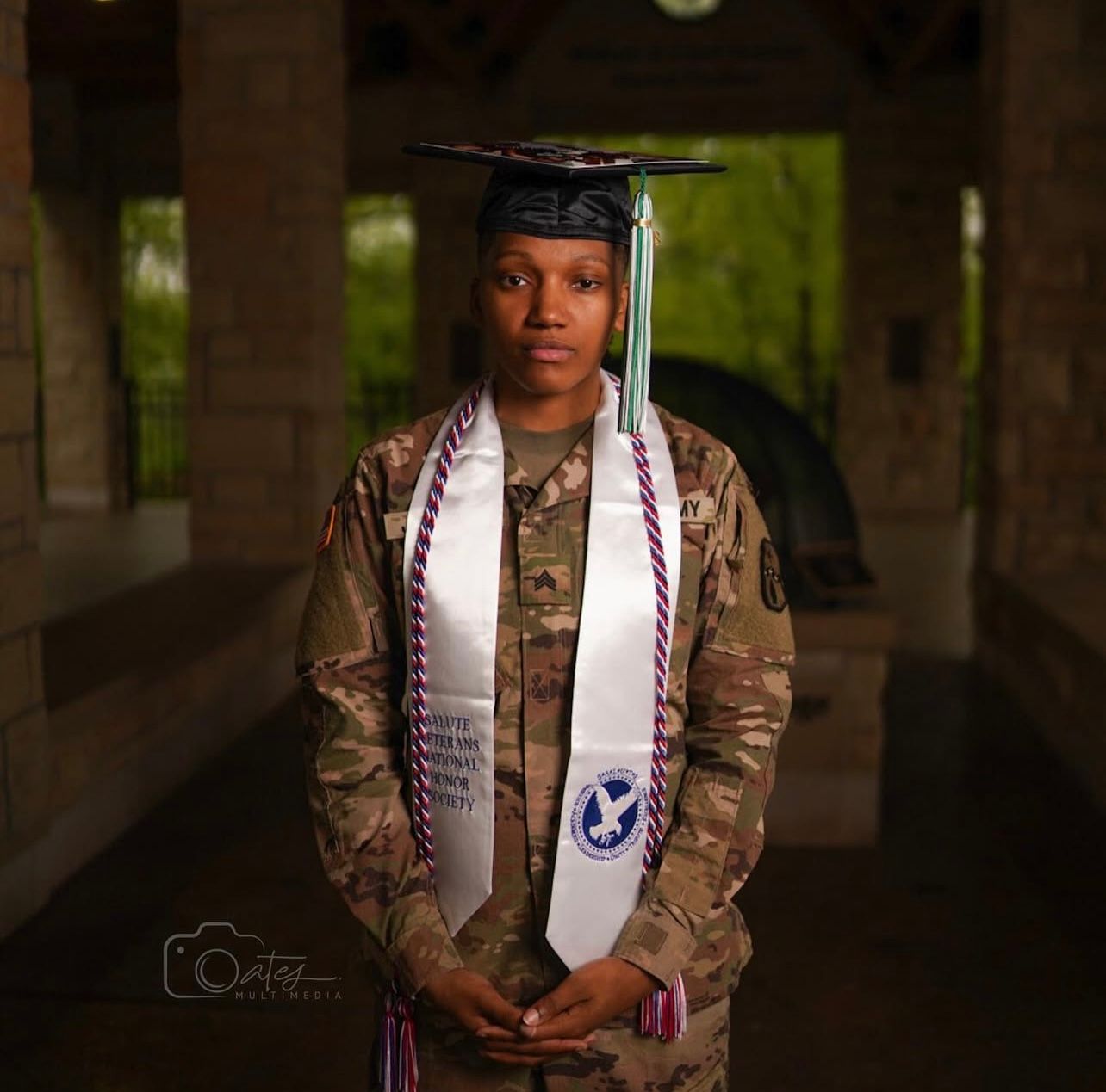 Military graduate in camouflage uniform with graduation stoles in dramatic lighting.